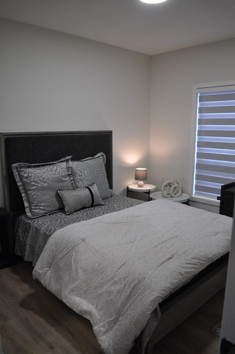 A bed with a grey comforter and pillows is in a room with a window and a lamp at The Laurent at Laurel Apartments, Edmonton, AB, T6T 2T5